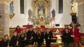 Eine Gruppe von Kindern steht in einer Kirche, hält Musikinstrumente, wahrscheinlich Klarinetten, vor einem geschmückten Altar und Statuen.