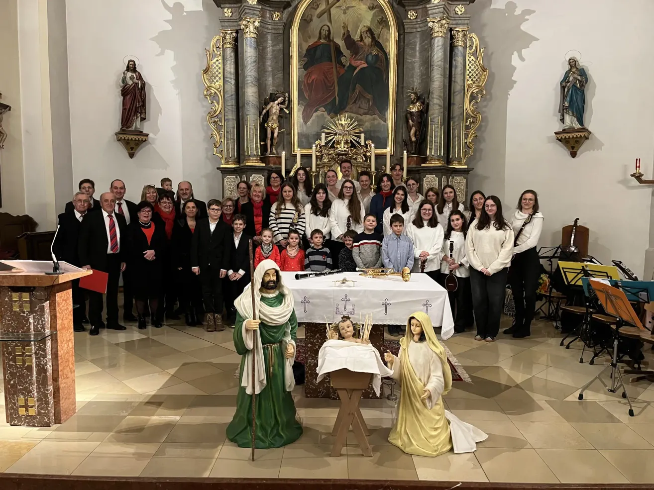 Ein Chor posiert für ein Gruppenfoto vor einem Kirchenaltar mit Krippenfiguren. Ein Tisch mit einem Kreuz und Musikinstrumenten steht im Vordergrund.