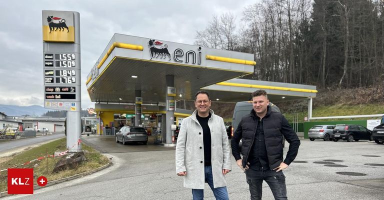 Zwei Männer stehen vor einer Tankstelle. Der linke trägt einen weißen Mantel und eine Brille, der andere einen schwarzen Mantel. Ein Auto ist an der Tankstelle geparkt.
