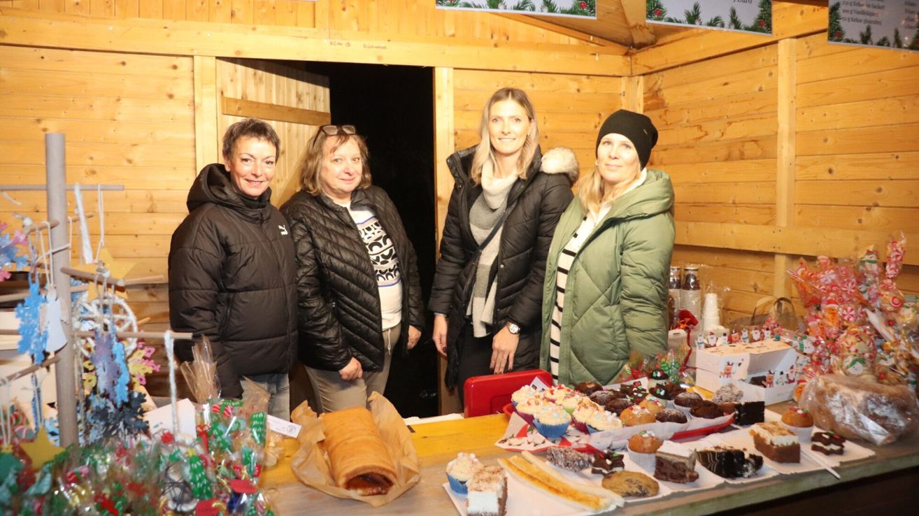Vier Frauen stehen in einer Holzhütte, lächelnd, mit einem Tisch voller Cupcakes und anderen Desserts vor ihnen. Die Hütte hat Holzwände und eine Tür hinten.
