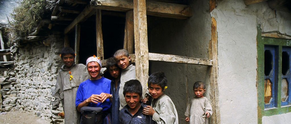 Eine Familie von sechs Personen steht vor einem rustikalen Stein- und Holzhaus. Die Frau lächelt, während die Kinder, die traditionelle Kleidung tragen, verschiedene Ausdrücke zeigen.