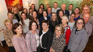 Eine Gruppe von Erwachsenen, wahrscheinlich ein Team, posiert für ein Foto. Sie tragen formelle Kleidung. Einige von ihnen tragen Brillen, Halsketten und Armbänder. Hinter ihnen befindet sich eine grüne Ziegelwand und eine Lampe. Links befindet sich ein Gemälde an der Wand.