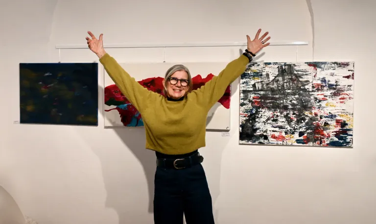 Eine Frau mit einer grünen Jacke und einer Brille steht mit ausgestreckten Armen vor einer weißen Wand mit zwei abstrakten Gemälden.