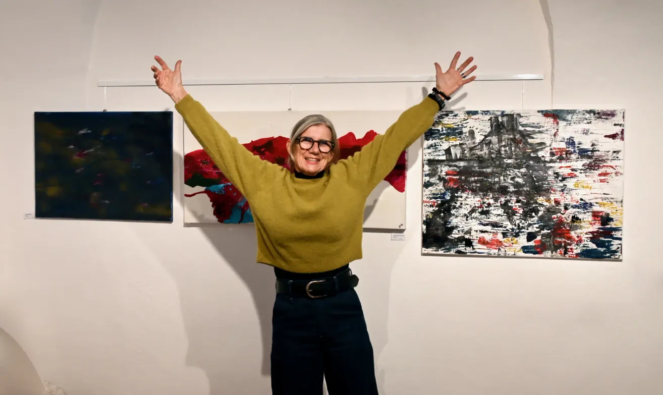 Eine Frau mit einer grünen Jacke und einer Brille steht mit ausgestreckten Armen vor einer weißen Wand mit zwei abstrakten Gemälden.