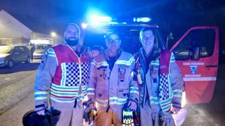 Drei Feuerwehrleute posieren für ein Foto vor einem Feuerwehrwagen bei Nacht. Sie tragen alle Sicherheitswesten. Der Mann auf der linken Seite hat einen Bart und lächelt. Der Mann in der Mitte trägt eine Brille. Der Mann auf der rechten Seite hält eine Taschenlampe. Der Feuerwehrwagen hat ein blaues Licht oben und den Text 'BREICHSDORF' auf der Vorderseite.