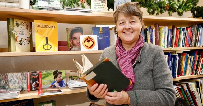 Eine Frau hält ein Buch in den Händen in einem mit Büchern und Dekorationen gefüllten Raum. Sie lächelt und steht vor einem Bücherregal.