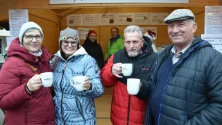 Eine Gruppe von Menschen in Winterkleidung lächelt und posiert für ein Foto. Sie halten weiße Tassen. Dahinter befindet sich ein Stand mit einem Schild, auf dem Super Huttenzuauber steht.