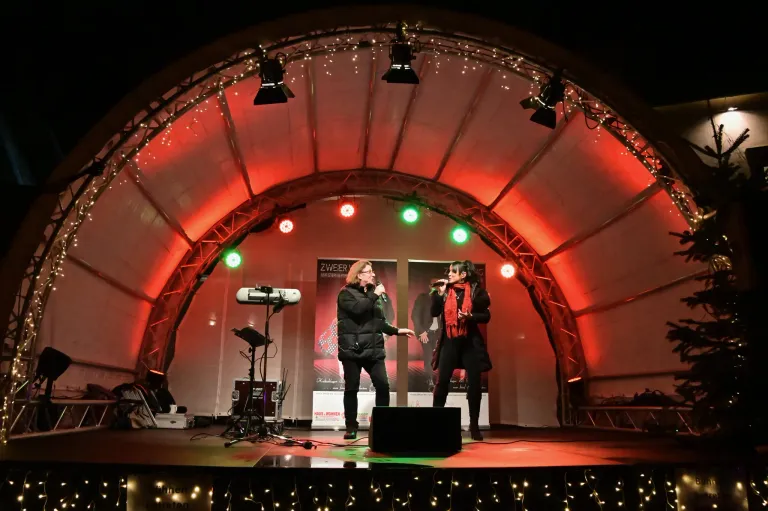 Zwei Frauen singen auf einer Bühne unter roten Lichtern. Ein Mikrofon und ein Lautsprecher sind auf der Bühne. Werbung steht hinter ihnen.