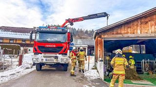 Ein roter Feuerwehrwagen mit dem Wort 'Feuerwehr' ist am Straßenrand geparkt. Drei Feuerwehrleute gehen auf die Scheune zu, einer hält ein Seil.