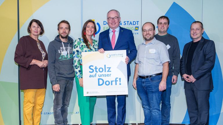 Fünf Personen stehen vor einem Hintergrund mit blau-weißem Design. Der Mann in der Mitte hält ein Schild mit der Aufschrift 'Stolz auf unser Dorf!' in Blau. Die Frau neben ihm trägt grüne Hosen. Hinter ihnen trägt ein weiterer Mann ein graues Sweatshirt.