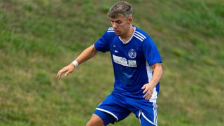 Ein Fußballspieler in einem blauen Trikot läuft auf einem Grasfeld. Er trägt ein weißes Armband und hat einen ernsten Gesichtsausdruck.