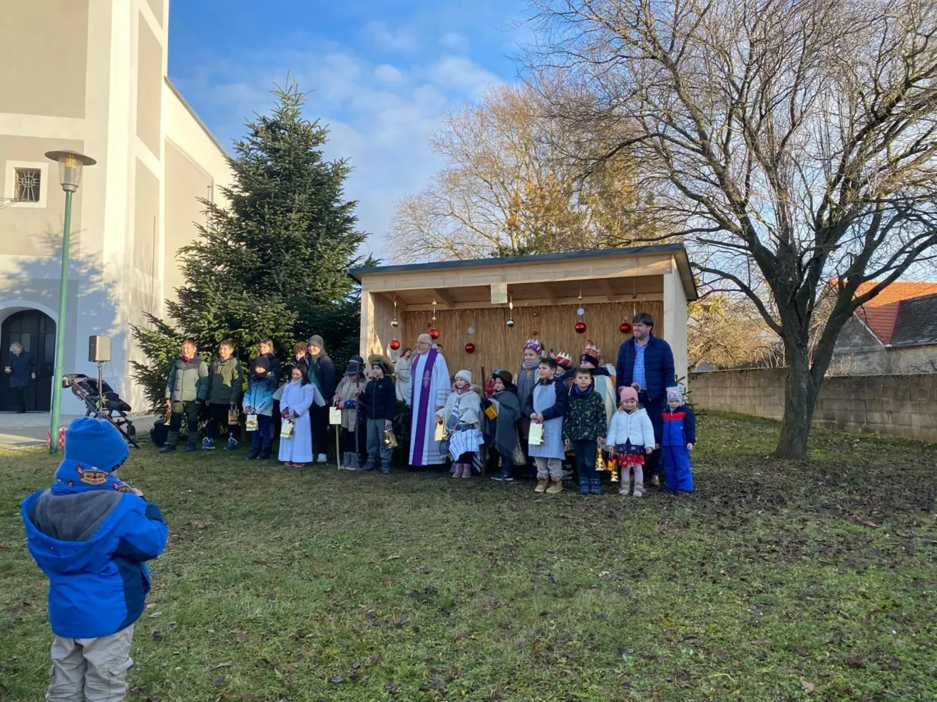 Eine Gruppe von Menschen steht vor einer Krippe im Garten. Sie sind in Winterkleidung gekleidet und einige halten Geschenke.