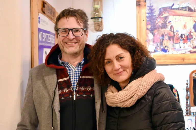 Ein Mann und eine Frau lächeln und stehen eng beieinander in einem Raum. Der Mann trägt eine Brille und ein gemustertes Hemd. Die Frau hat lockiges Haar und trägt einen Schal. Hinter ihnen ist eine Wand mit einem Holzrahmen und einem Poster.
