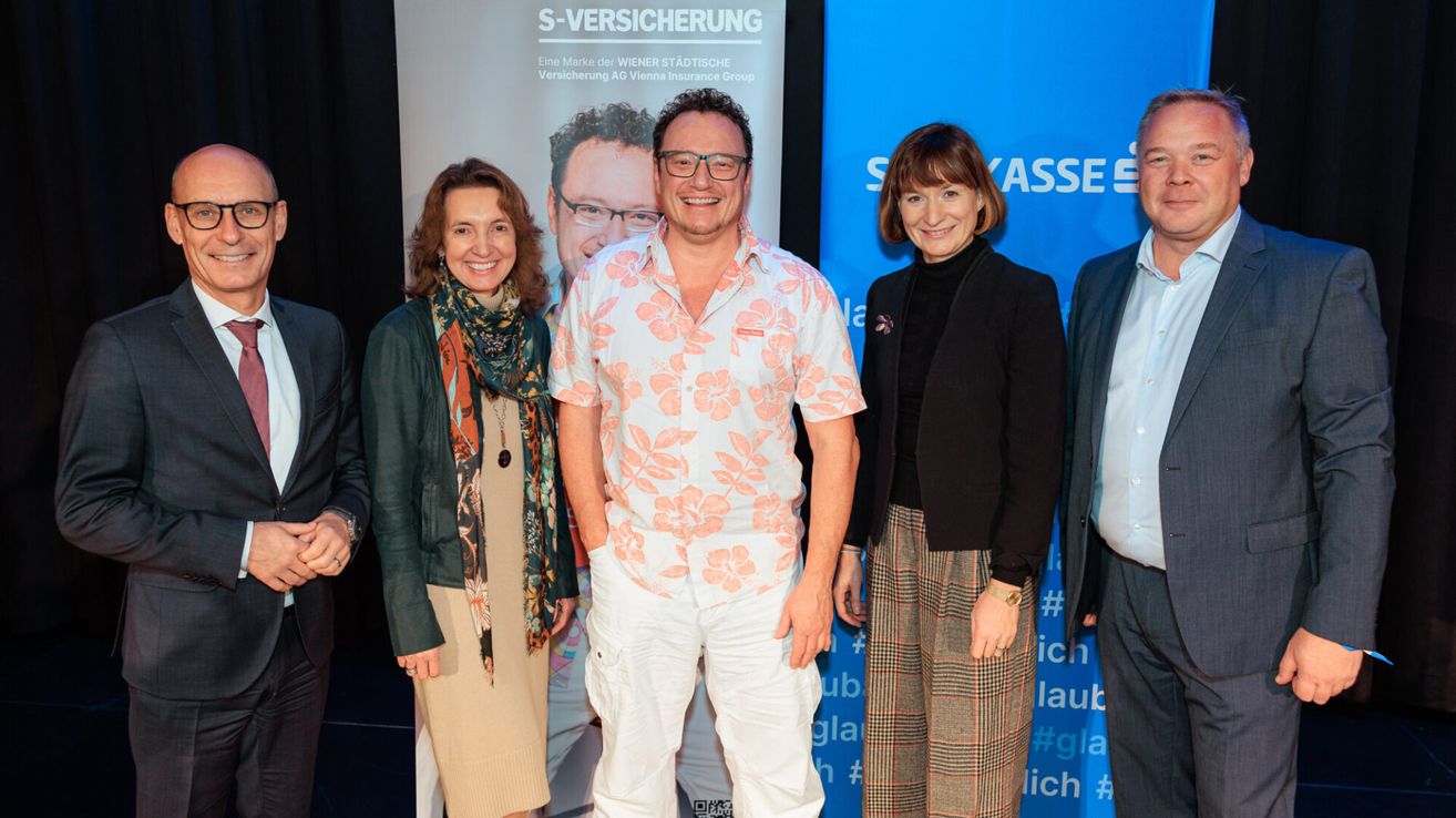 Vier Personen stehen lächelnd vor einem blauen Banner mit den Worten 'S-Versicherung' und einem Logo. Der Mann in der Mitte trägt eine Brille und ein Blumenhemd, während die Frauen Schals und Jacken tragen.