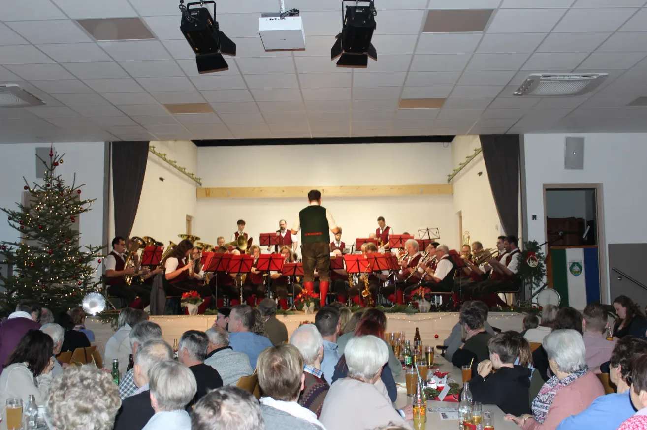 Eine Band spielt auf der Bühne, während das Publikum von vorne zusieht. Die Bühne ist mit Blumen und Lichtern geschmückt. Das Publikum sitzt auf Stühlen, und einige stehen. Sie genießen Getränke, während sie die Aufführung verfolgen.