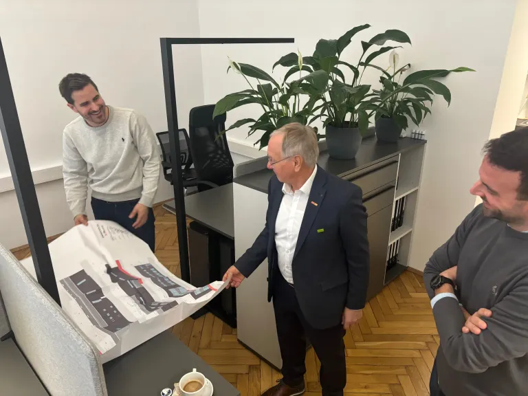 Zwei Männer in Anzügen betrachten ein großes Blatt Papier. Hinter ihnen lächelt ein Mann. Im Hintergrund stehen Topfpflanzen auf einem Schrank und einem Schreibtisch.