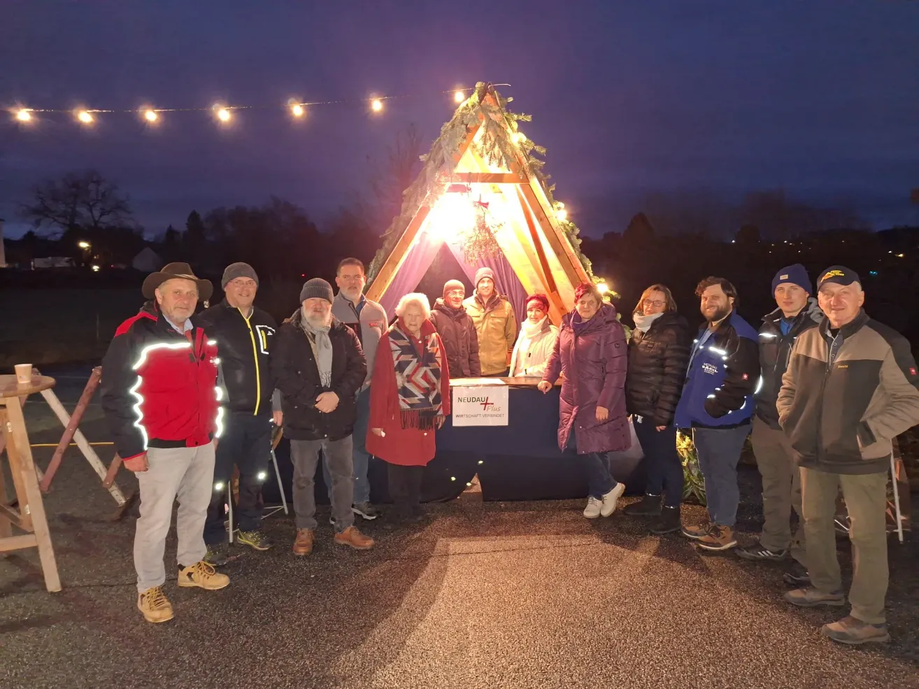 Eine Gruppe von Menschen steht vor einem geschmückten Zelt mit Lichterketten und einem Tisch mit einem Schild. Sie sind warm gekleidet für das kalte Wetter.