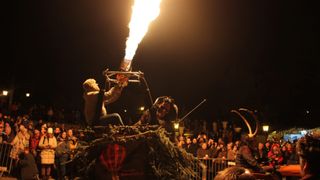 Ein Heißluftballon wird nachts zur Abflugvorbereitung gestartet, eine Person sitzt im Korb und ein Feuer speiender Künstler in der Nähe, eine Menge beobachtet.