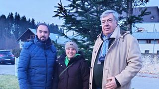 Drei Personen stehen lächelnd vor einem Weihnachtsbaum. Ein Mann links trägt einen blauen Mantel, eine Frau in der Mitte trägt eine Mütze und ein grünes Shirt, und ein Mann rechts hat einen blauen Schal.