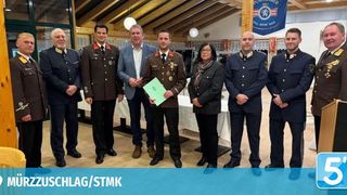 Eine Gruppe von Militäroffizieren in Uniformen posiert für ein Foto in einer Halle. Eine Frau mit Brille und Schal steht in der Mitte. Ein Mann in Anzug hält eine grüne Mappe. Hinter ihnen steht ein Tisch mit weißer Tischdecke.