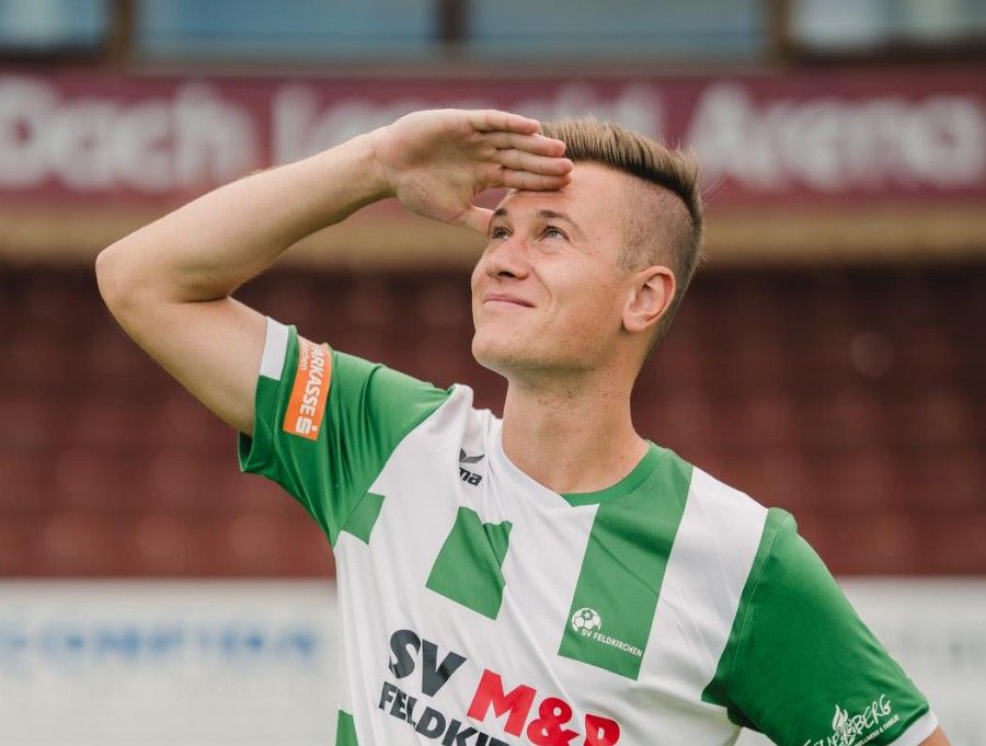 Ein Fußballspieler in einem grün-weißen Trikot mit SV Feldkirchen-Logo salutiert auf einem Feld.