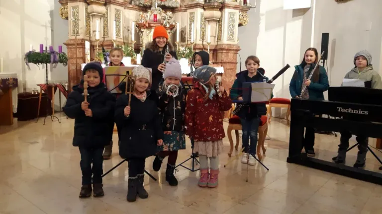 Eine Gruppe von Kindern steht in einer Kirche und hält Musikinstrumente. Die Frau in der Mitte hält eine Klarinette. Vor ihnen steht ein Notenständer.