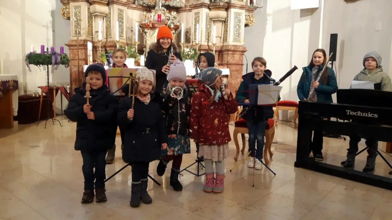 Eine Gruppe von Kindern steht in einer Kirche und hält Musikinstrumente. Die Frau in der Mitte hält eine Klarinette. Vor ihnen steht ein Notenständer.