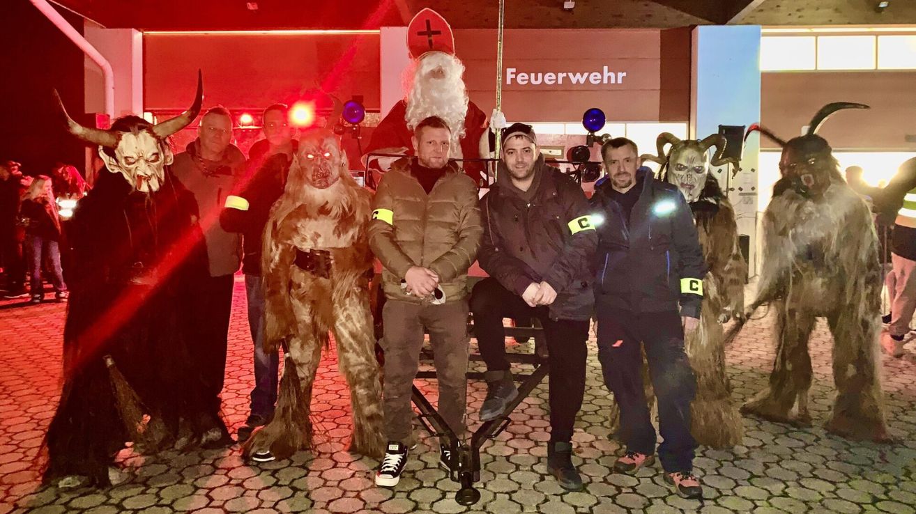 Eine Gruppe von Menschen, darunter zwei in Kostümen, posiert für ein Foto vor einem Feuerwehrhaus mit einer Weihnachtsmannfigur im Hintergrund.