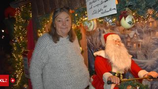 Eine Frau in einem grauen Pullover posiert neben einer Weihnachtsmann-Figur in einem festlich geschmückten Raum. Hinter ihr hängt ein weißes Schild mit schwarzen Schriftzügen von einer Weihnachtsgirlande.