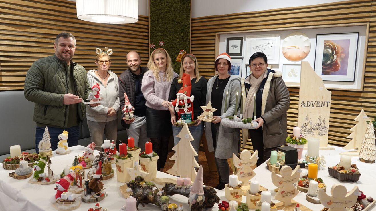A group of people stand in a room decorated for Christmas, each holding festive items. A table in front of them is laden with candles, ornaments, and Christmas tree figures. Behind them, a wall adorned with various framed pictures creates a festive backdrop.