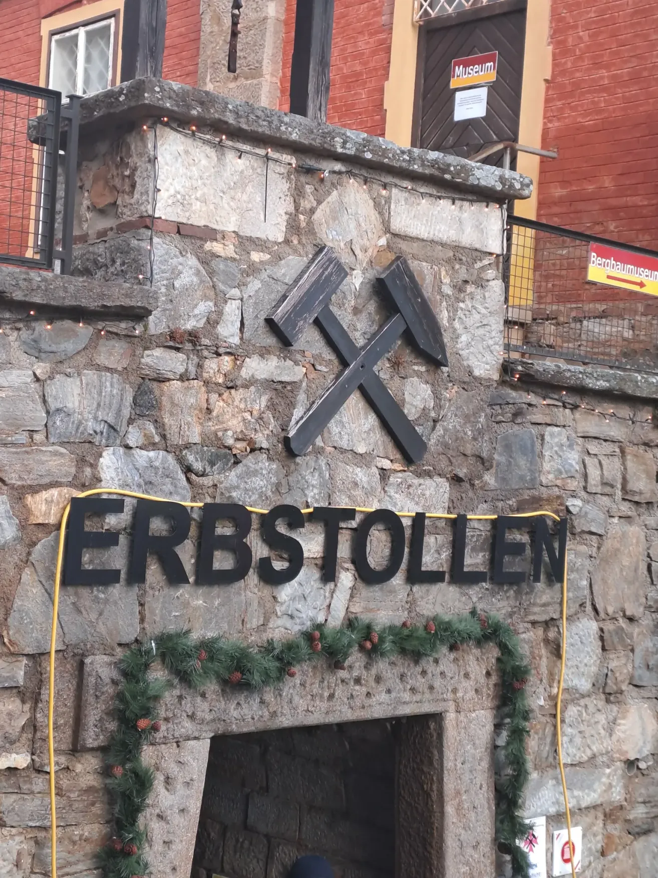 Ein Steingebäude mit einem Schild, das 'ERBSTOLLEN' steht. Darauf sind zwei gekreuzte Spitzhacken montiert. Unten schmückt eine festliche Girlande mit Tannenzapfen.