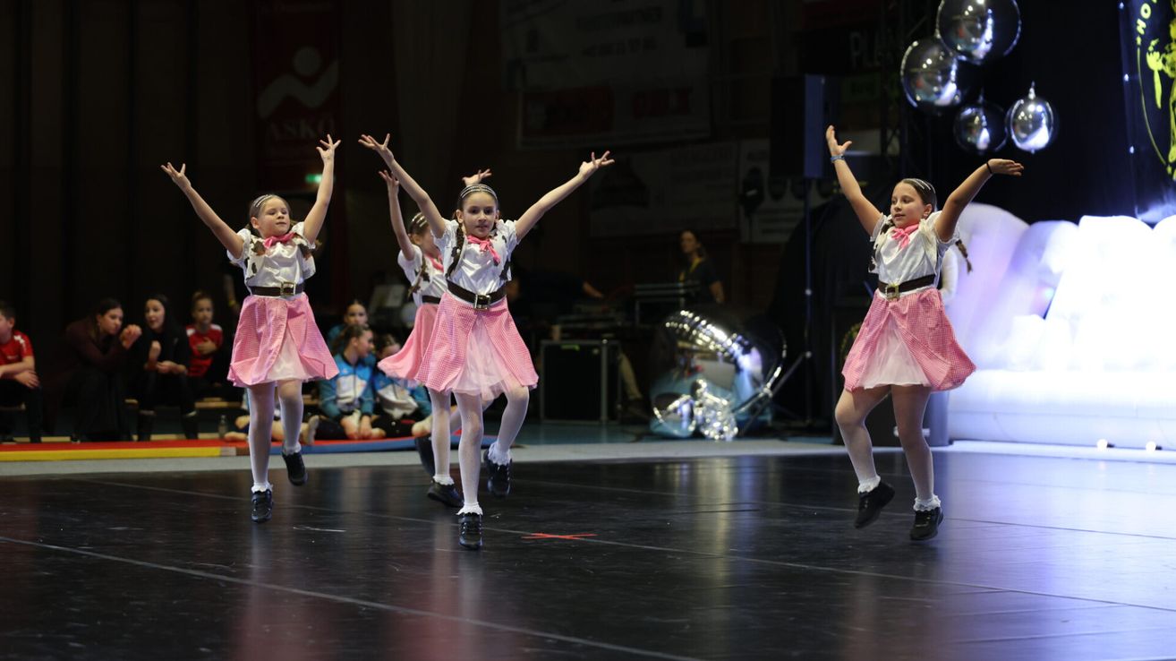 Mehrere junge Mädchen in rosa Kleidern und schwarzen Schuhen führen eine synchronisierte Tanzaufführung auf der Bühne mit erhobenen Armen durch. Dahinter sind teilweise ein Schlagzeug und eine Frau zu sehen.