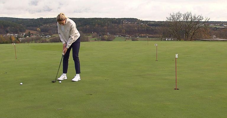 Eine Golfspielerin ist auf einem Golfplatz, hält einen Schläger und ist dabei, einen Ball in Richtung eines Lochs zu schlagen. Der Platz ist grün und mehrere Flaggen markieren die Löcher.