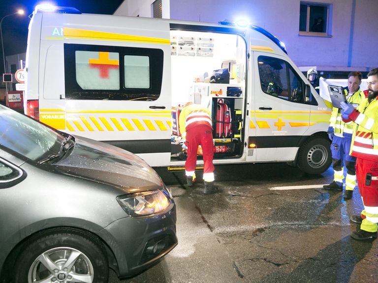 Ein Krankenwagen ist auf einer nassen Straße geparkt, seine Lichter sind an. Eine Person in einem gelben und roten Sicherheitsanzug lädt Ausrüstung in den Krankenwagen.