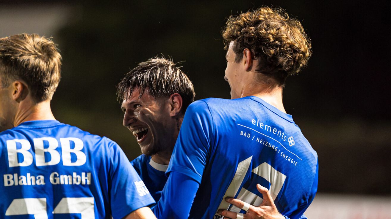 Drei Fußballspieler in blauen Trikots feiern gemeinsam. Der Spieler auf der rechten Seite trägt ein Trikot mit 'elements' und 'Bad Heizung Energie' auf dem Rücken.