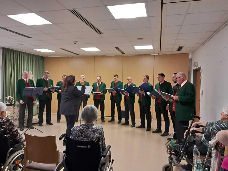 Ein Chor in grünen Westen singt in einem Raum mit einer Holzwand. Eine Frau im Rollstuhl schaut zu.