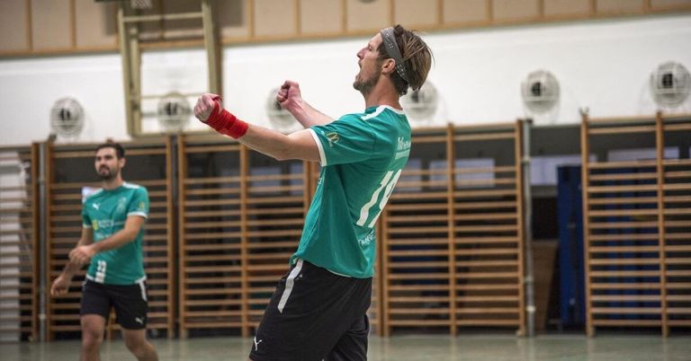 A man in a green shirt with the number 11 on it is celebrating in an indoor sports facility, wearing a red headband and red armband.