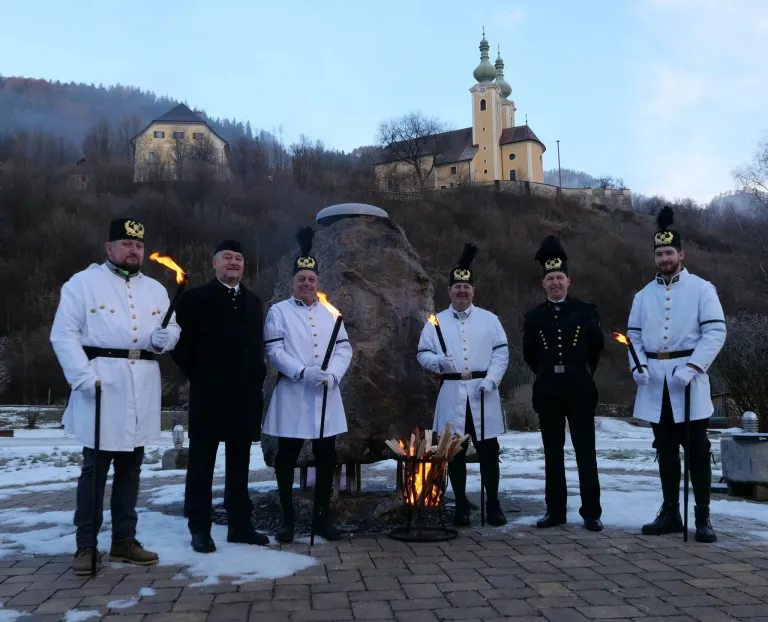 Sechs Männer in Militäruniformen, mit Fackeln, stehen vor einem Lagerfeuer mit einem großen Felsen im Hintergrund. Eine Kirche ist auf einem Hügel sichtbar.