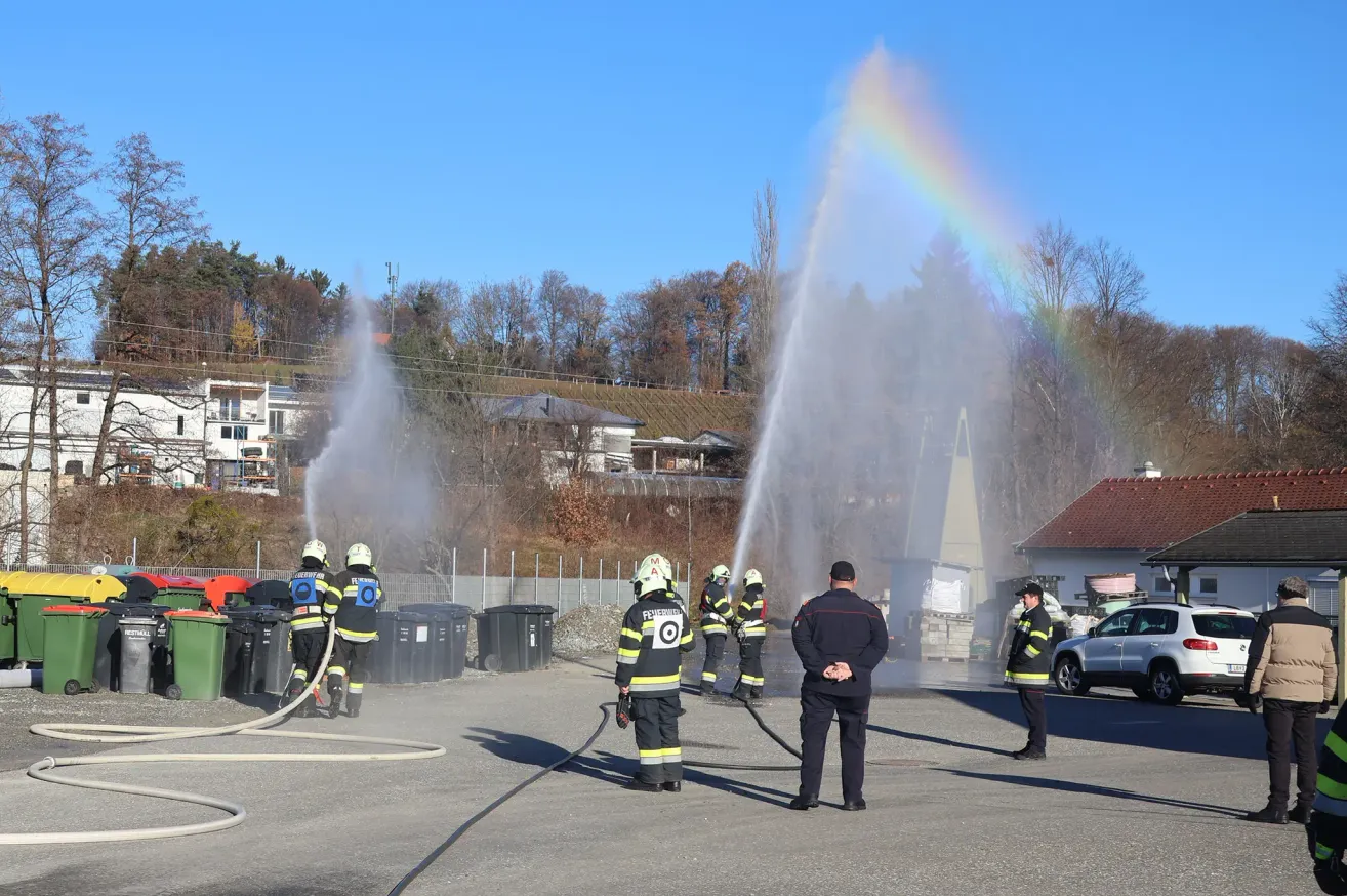 Feuerwehrleute üben mit einem Feuerwehrschlauch in einem Außenbereich mit einem Regenbogen am Himmel, umgeben von Bäumen und Gebäuden.