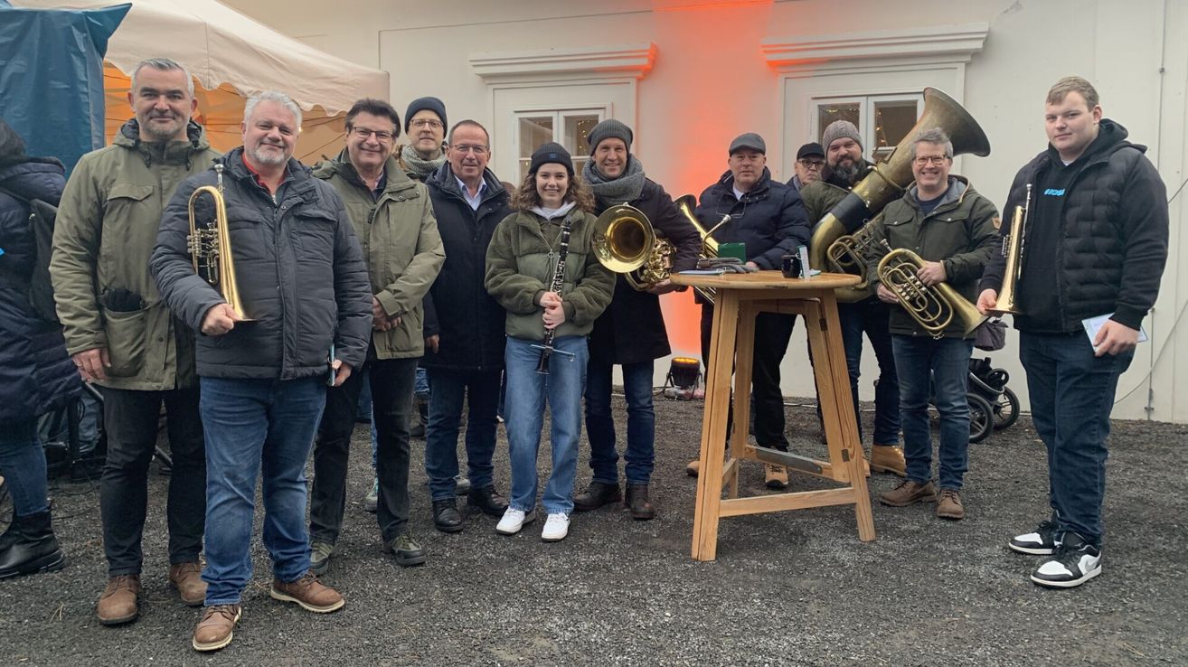 Eine Gruppe von Menschen, einige in Winterkleidung, posiert für ein Foto vor einem Gebäude. Eine Frau in der Mitte hält eine Klarinette. Musikinstrumente wie Tuben stehen auf einem Holztisch.