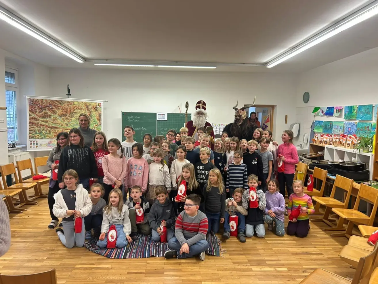 Gruppenfoto von Schülern in einem Klassenzimmer mit dem Weihnachtsmann und Krampus, beide in Kostümen. Schüler halten rote Tüten, möglicherweise Geschenke. Ein Tafel und Dekorationen im Hintergrund.