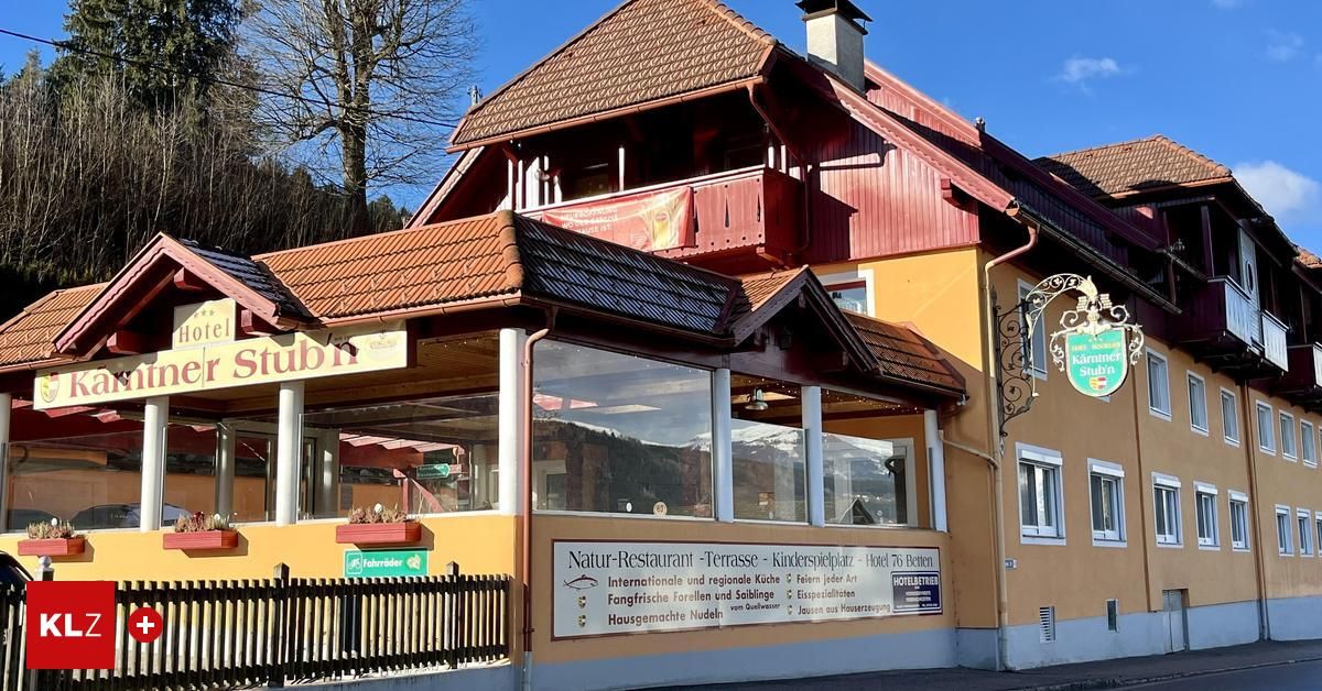 Ein gelbes Gebäude mit braunen Dächern, einer Terrasse und großen Fenstern. Es hat ein Schild mit der Aufschrift 'Natur-Restaurant Terrasse Kinderspielplatz Hotel 76 Betten'. Vor dem Gebäude steht ein Zaun und es gibt Pflanzen. Im Hintergrund befinden sich ein Baum und Berge.