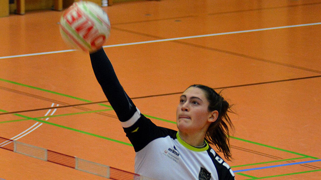 A female volleyball player is throwing a ball with her left hand. She is wearing a white and black jersey. The court has orange and green markings.