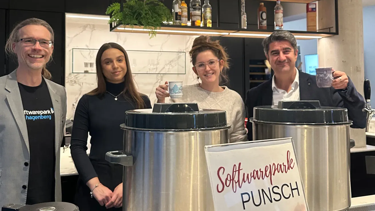 Drei Personen, zwei Frauen und ein Mann, lächeln und stehen vor einem großen Metalltopf. Die Frau auf der linken Seite trägt ein schwarzes Oberteil und eine Halskette. Die Frau in der Mitte hält einen Becher. Dahinter befinden sich Regale mit Likörflaschen.