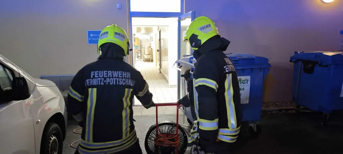 Two firefighters in yellow helmets are entering a building, one holding a tool cart. Behind them, a door is open, leading to a hallway with a white floor.