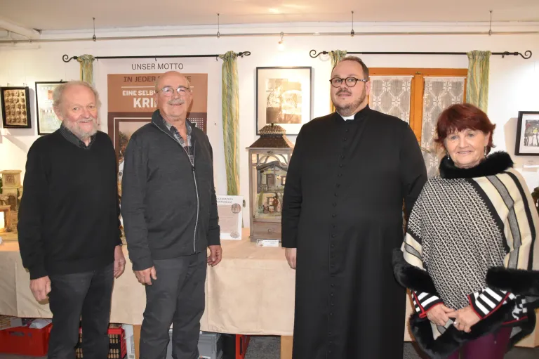 Vier Personen stehen in einem Raum mit einem Tisch, einer in einem schwarzen Gewand, lächelnd, und drei andere mit verschiedenen Ausdrücken. Dahinter befindet sich ein Banner und ein Bilderrahmen.