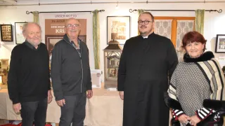 Vier Personen stehen in einem Raum mit einem Tisch, einer in einem schwarzen Gewand, lächelnd, und drei andere mit verschiedenen Ausdrücken. Dahinter befindet sich ein Banner und ein Bilderrahmen.