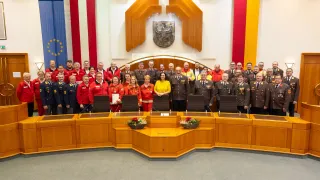 Eine Gruppe uniformierter Personen steht in einem Konferenzraum mit einer Frau, die ein Dokument in der Mitte hält. Sie sind von Flaggen und Emblemen umgeben.
