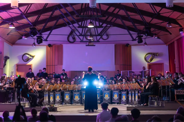 Ein Orchester spielt auf einer Bühne unter lila Beleuchtung, wobei der Dirigent führt. Das Publikum sitzt vor der Bühne und beobachtet die Aufführung.