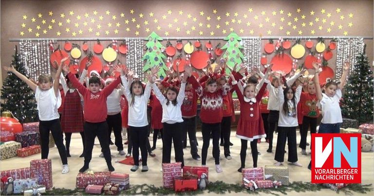 Eine Gruppe von Kindern in Weihnachtskostümen, mit roten Pullovern und Stirnbändern, führt einen Weihnachtstanz vor einem festlichen Hintergrund auf.